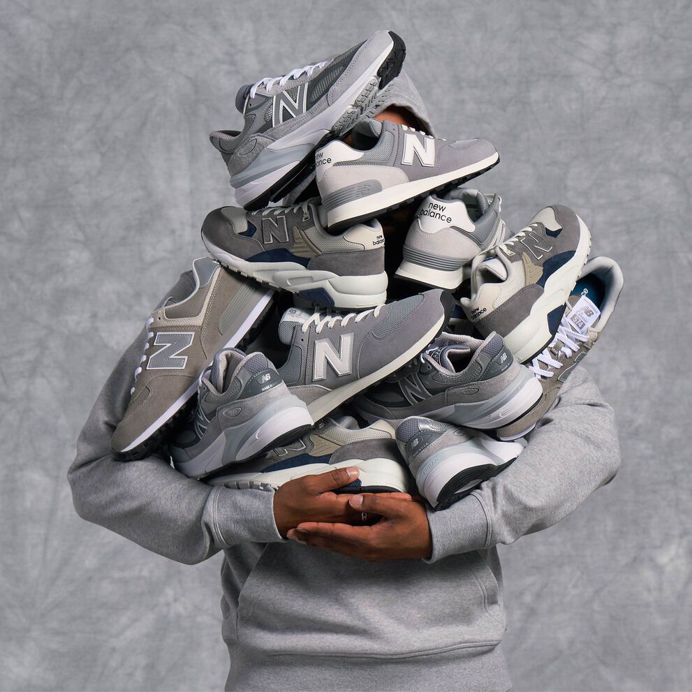 A person wearing a grey New Balance hoodie, holding a pile of grey New Balance shoes in their arms with their face obscured.