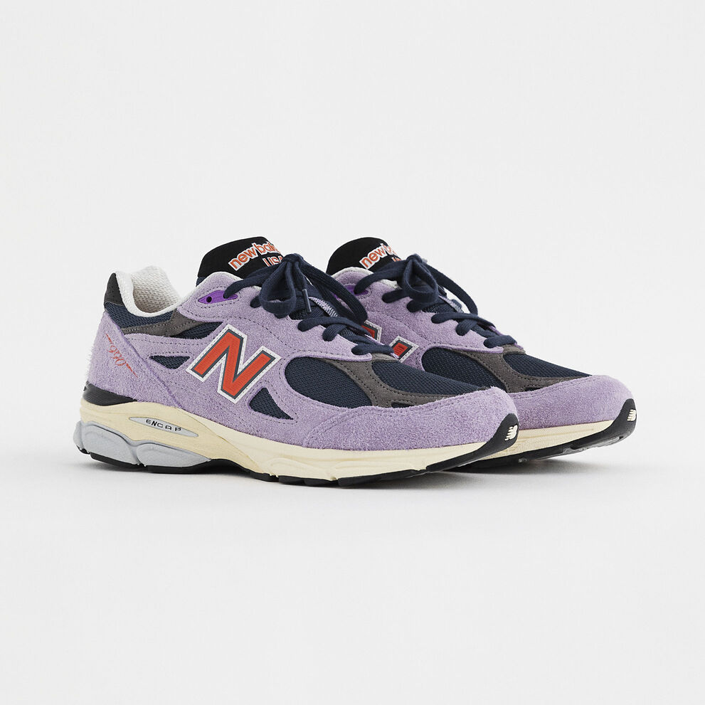 A pair of lavender 990v3 sneakers on a grey studio environment.