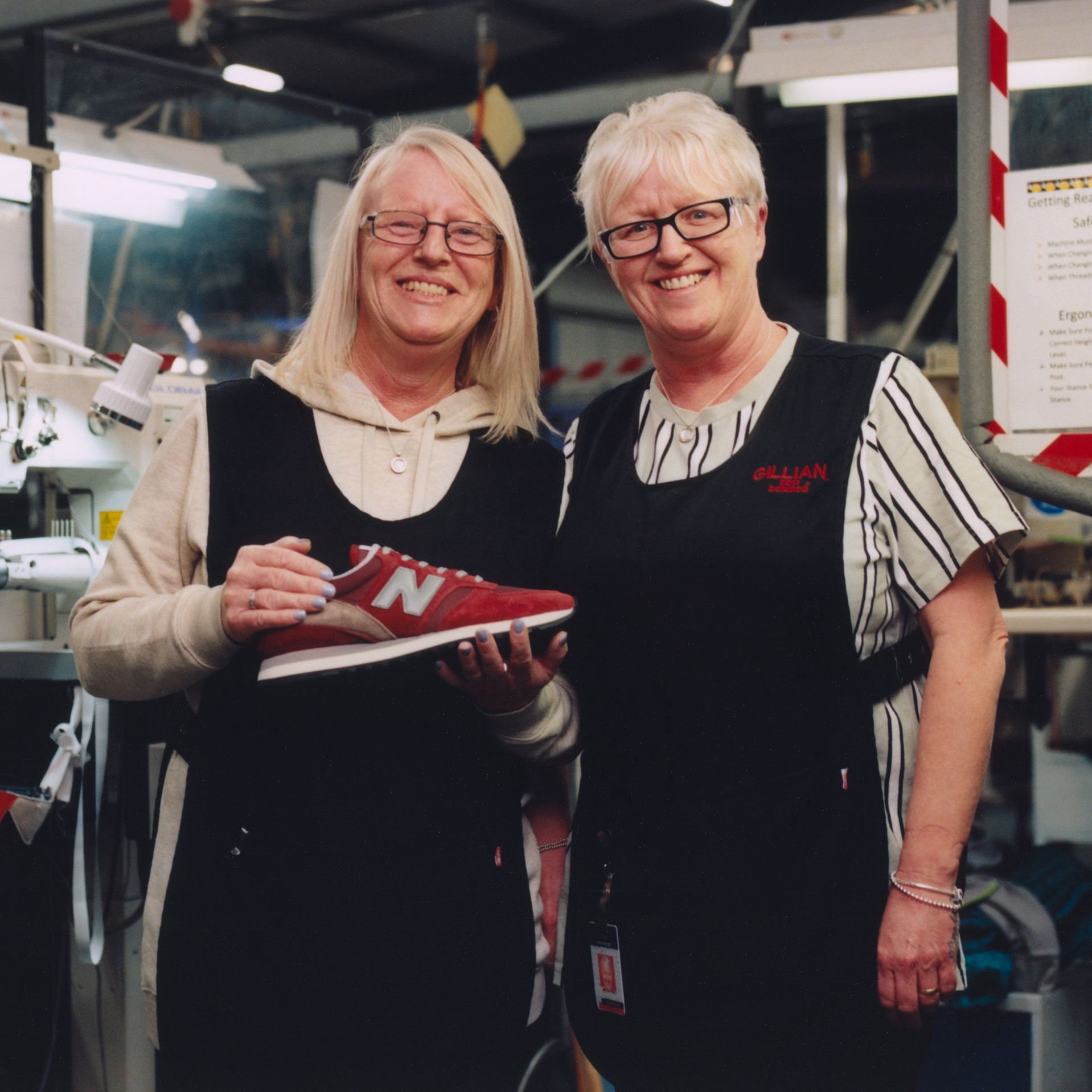 Two female workers at the Flimby factory standing together smiling at the camera with one holding a red Made in UK shoe.